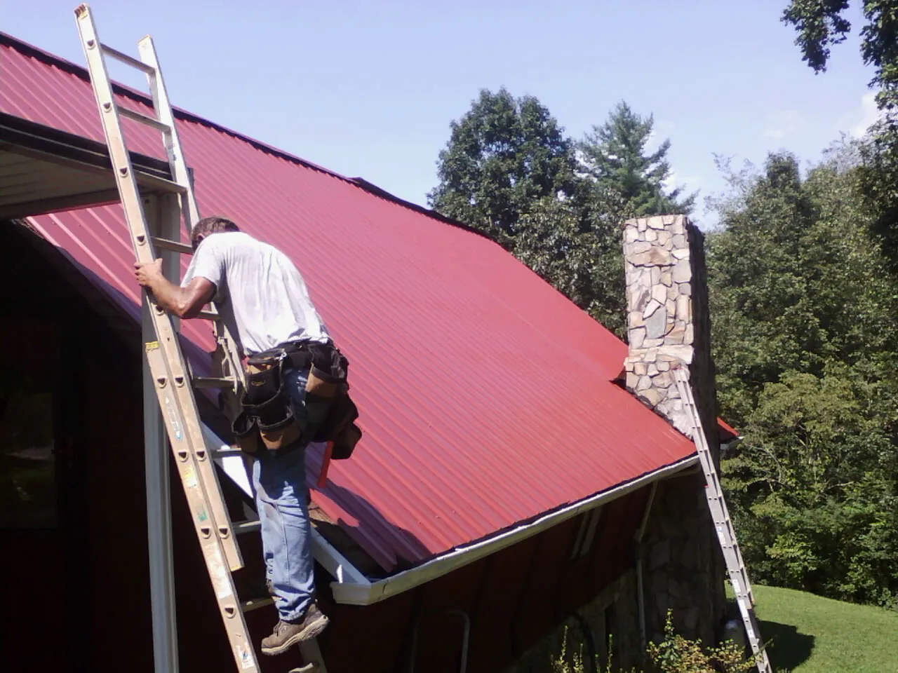 Roofer on a ladder beside a red metal roof and stone chimney — Soffit & Fascia Repair by Roofing Brothers in Hamtramck
